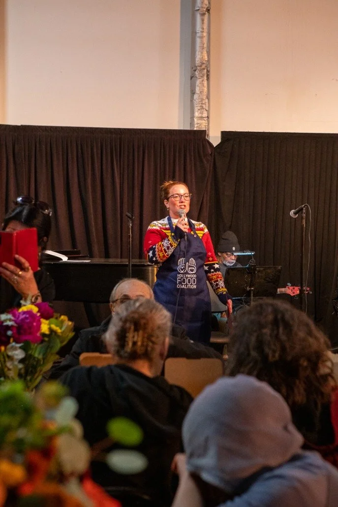 A woman in a festive sweater and apron singing into a microphone on stage, with two musicians and an audience in the foreground at a community event.