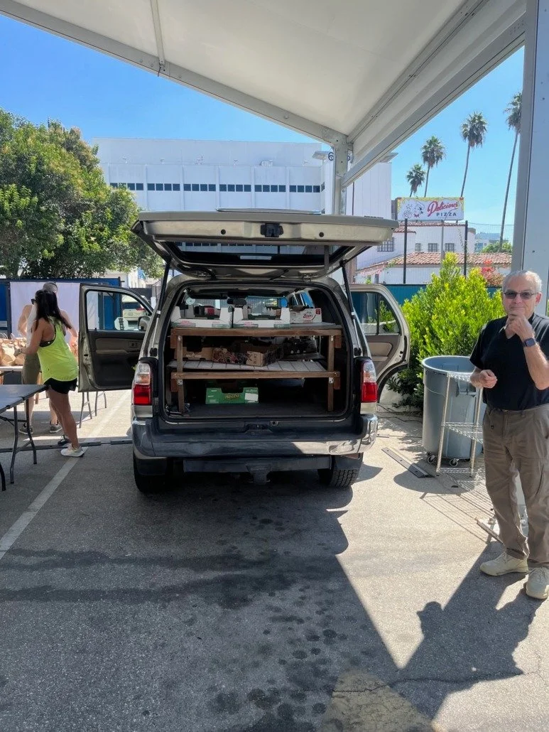 A gray vehicle with its rear hatch open, parked under a canopy at an outdoor setting with a man standing to the right wearing sunglasses, a black shirt, and beige pants, and a woman on the left in a yellow top and black shorts.
