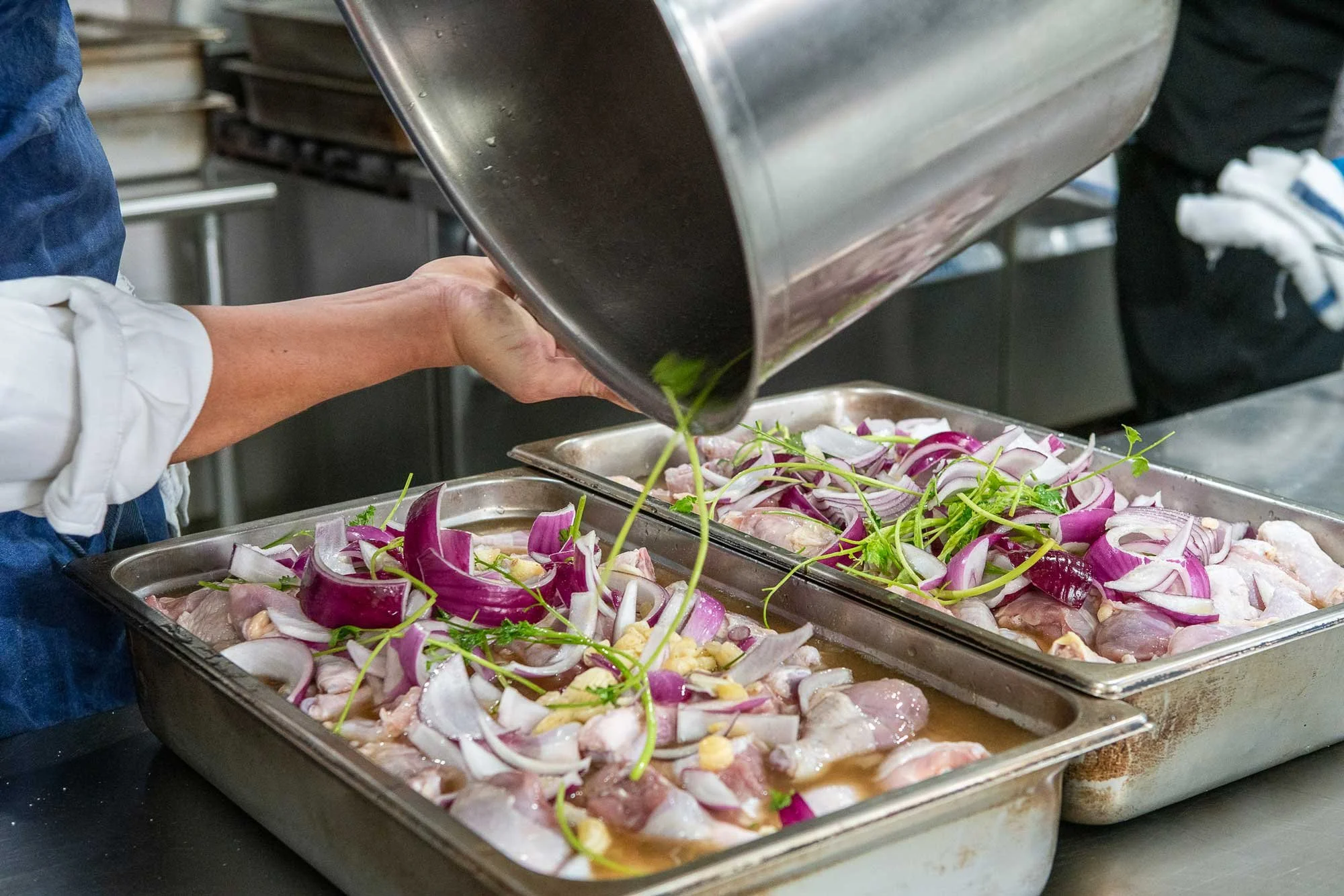 Someone pouring a liquid over seasoned raw chicken with sliced red onions and herbs in metal trays in a commercial kitchen.