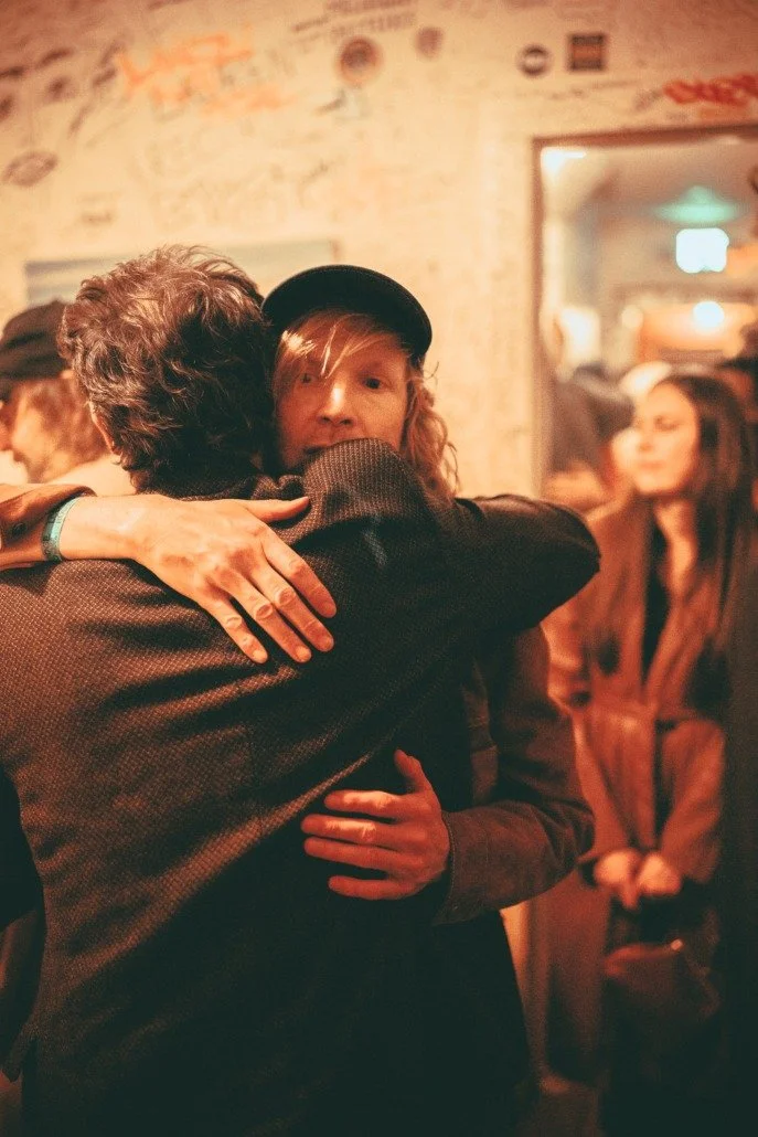 Two people hugging at a social gathering in a warm, dimly lit room with graffiti on the wall, and a woman sitting in the background observing.