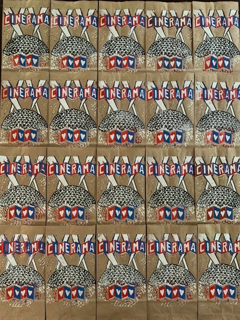 Multiple paper bags with a repeated graphic of a cinema sign, an umbrella, and a building with hearts, with the word 'CINERAMA' at the top on each bag.