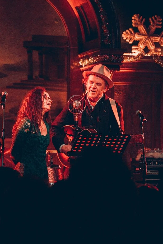 Two musicians perform on stage with microphones, one singing and the other playing guitar, under Christmas decorations including a large snowflake ornament.