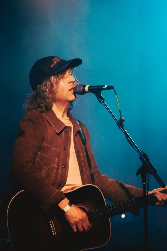 A person singing into a microphone and playing an acoustic guitar on stage, wearing a dark cap and brown jacket, with blue lighting in the background.