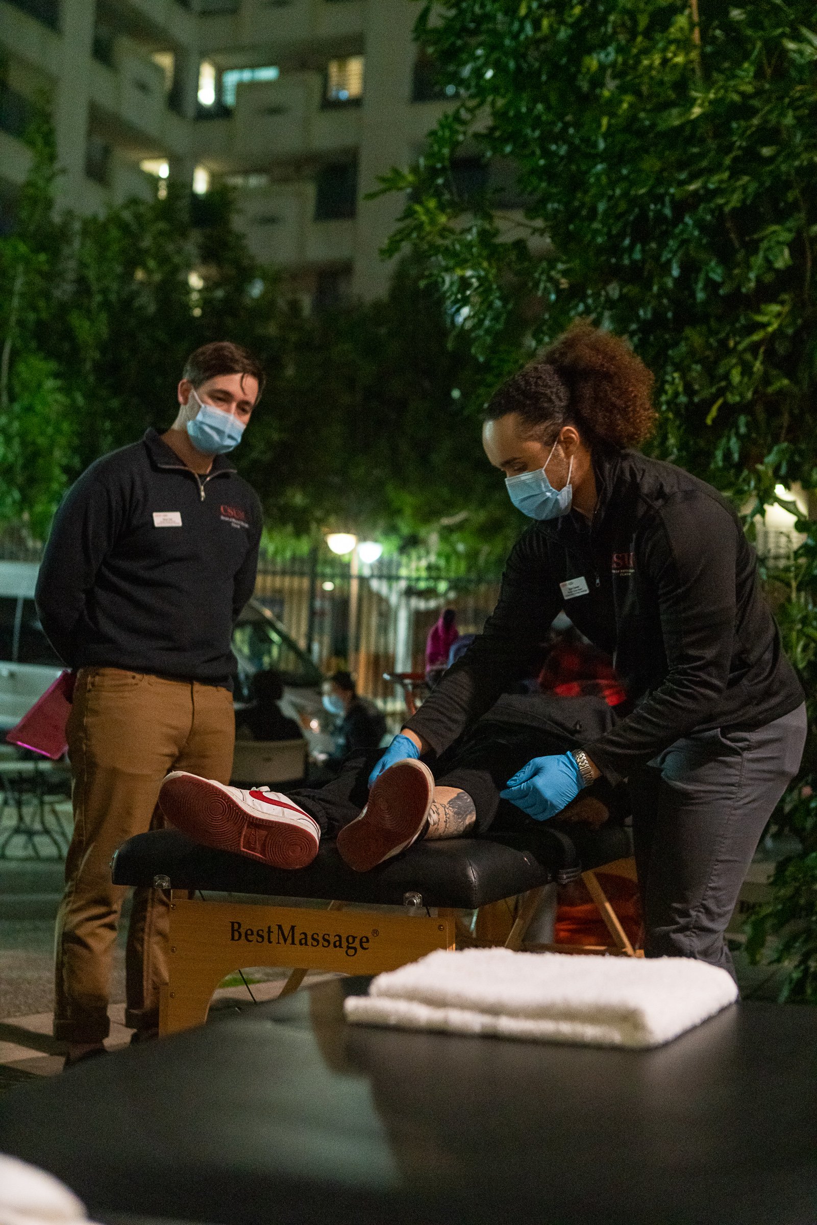 Two massage therapists performing a massage on a client lying face down on a massage table outdoors at night. All three individuals are wearing masks.