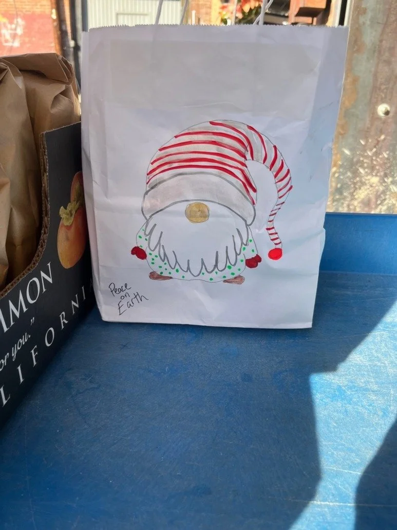 A white paper gift bag decorated with a drawing of a gnome or elf wearing a red and white striped hat, with the words "Peace on Earth" written in the bottom left corner.