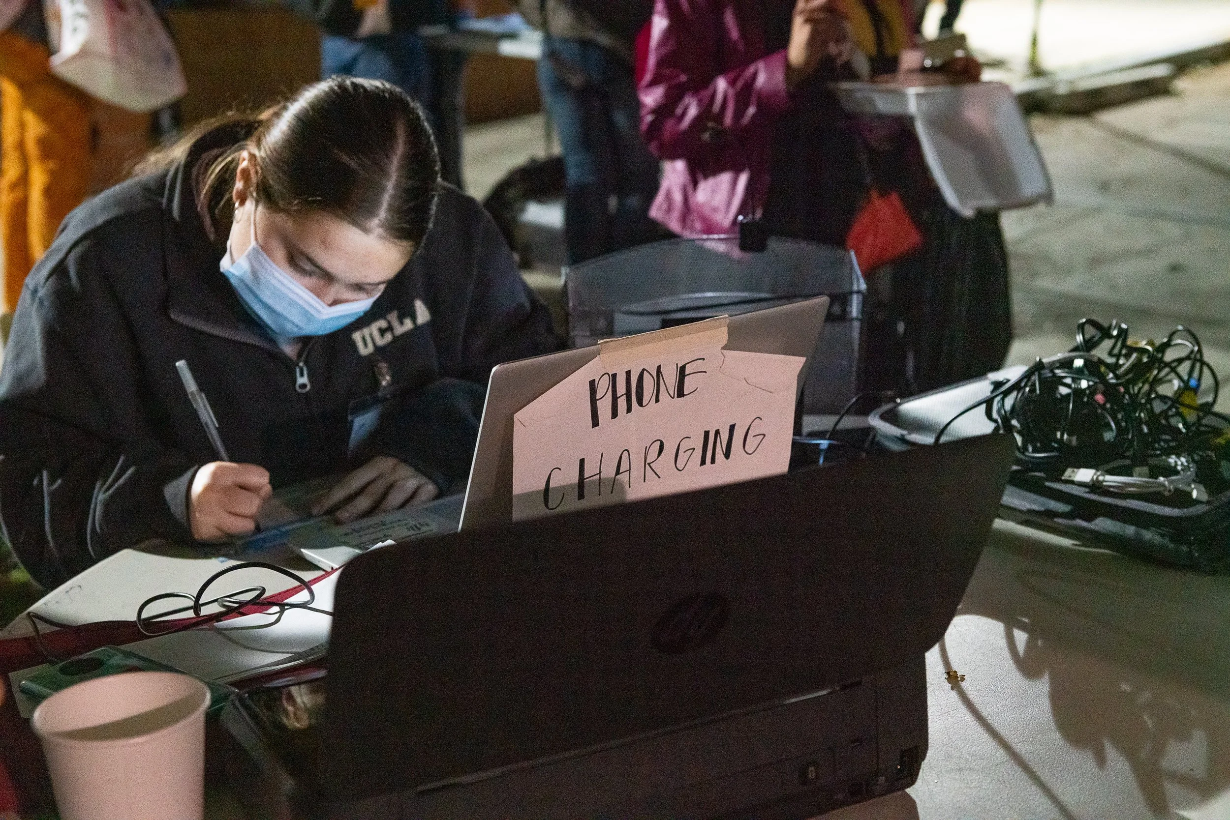 A woman wearing a face mask charging her phone at a table with various electronic equipment, with a sign that says 'PHONE CHARGING' taped to her laptop.