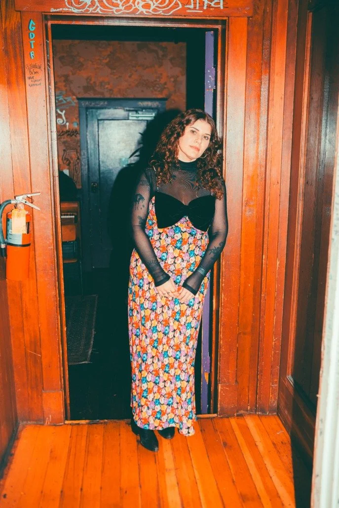 A woman with curly hair wearing a black mesh top with long sleeves and a floral maxi dress, standing in a doorway of a wooden room.