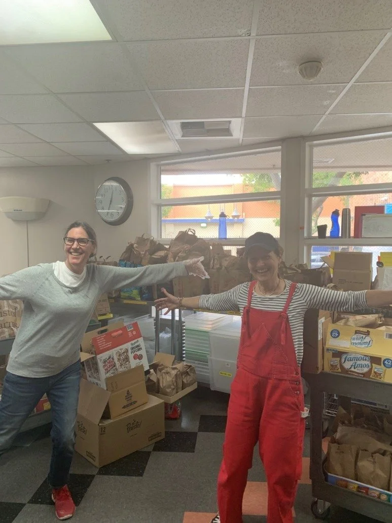 Two women smiling and extending their arms in a room filled with boxes and bags, likely a food bank or community service location.