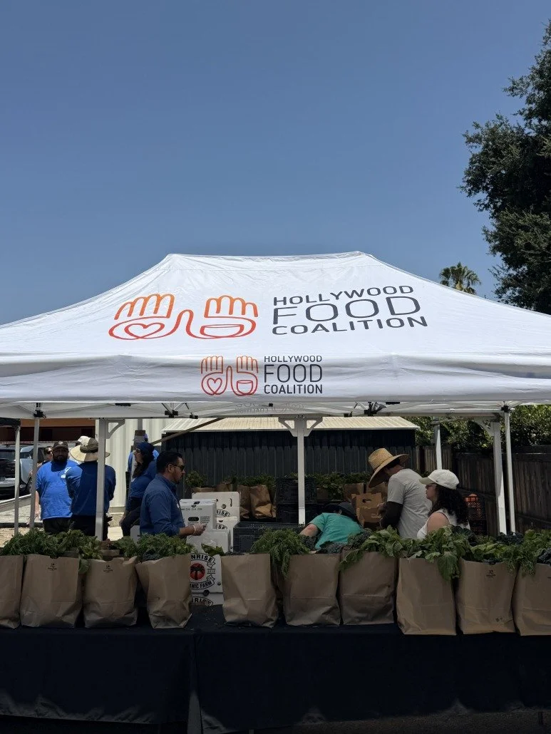 A white canopy tent with the logo and name of the Hollywood Food Coalition, featuring two hands making a heart symbol. Several people are gathered around a table with brown paper bags filled with vegetables, likely at a community food distribution ev