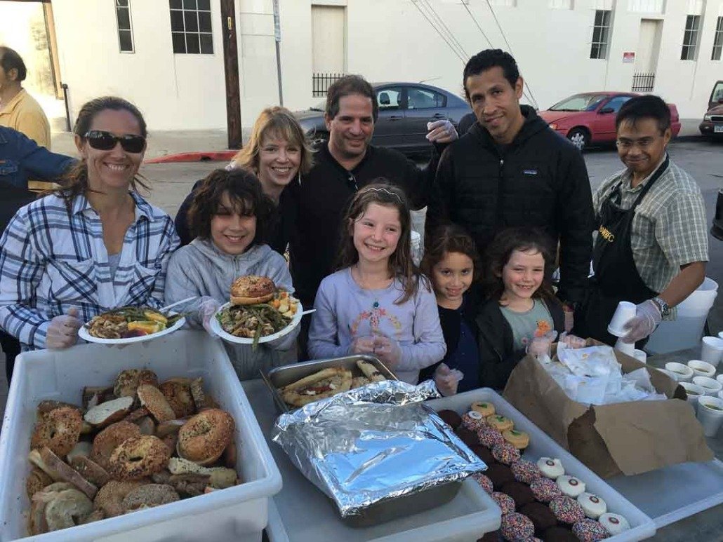 Group of people serving and enjoying food outdoors, with trays of baked goods, hot dishes, and decorated cookies on a table.