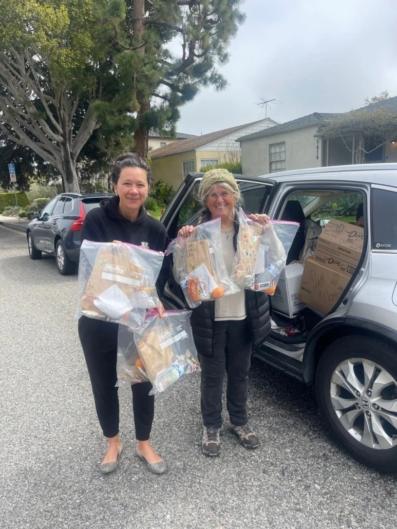 Two women standing beside a car with open trunk, holding large ziplock bags filled with groceries and supplies, parked on a residential street with trees and houses in the background.