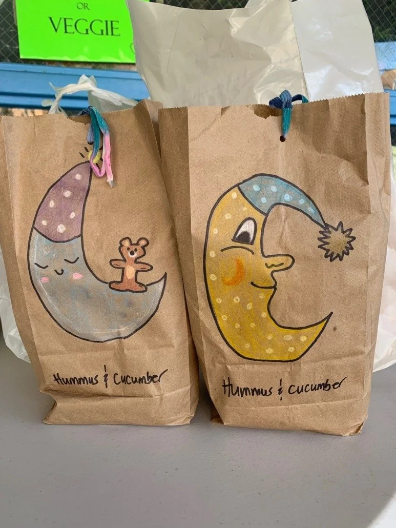Two brown paper bags with hand-drawn illustrations of a moon and a smiling face wearing a nightcap, one with a bear and the other with a face, both labeled "Hummus & Cucumber."