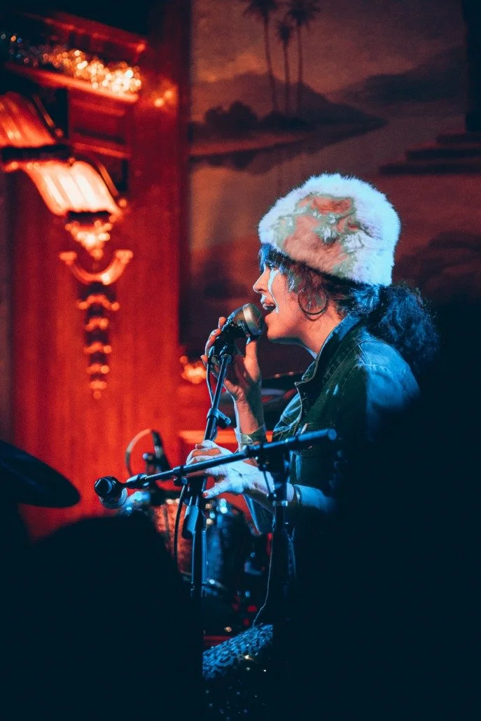 A woman singing into a microphone, wearing a white fuzzy hat with a colorful design, in a cozy indoor setting with warm lighting and wooden decor.