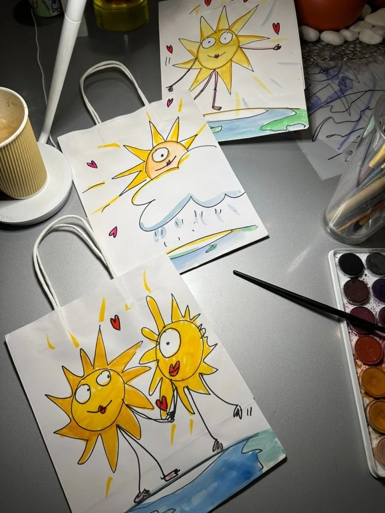Three illustrated paper bags with cartoon sun characters on a table, labeled as matching the drawn artwork. The artwork features cheerful suns with expressive faces and limbs, drawn with bright colors and surrounded by small hearts and backgrounds of
