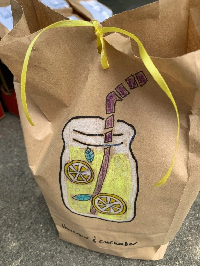 A paper bag with a hand-drawn illustration of a mason jar containing a lemon and cucumber infused water with lemon slices, cucumber slices, and leaves, tied with a yellow ribbon. The text "Lemon & cucumber" is written at the bottom.