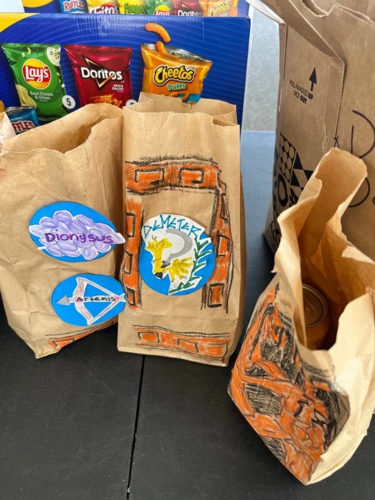 Three paper bags decorated with drawings and stickers, placed on a table with snack bags in the background. The middle bag has a drawing of a brick wall around a blue circle with a yellow creature and the text 'DЕМЕРСИЯ?' around it. The left bag has 
