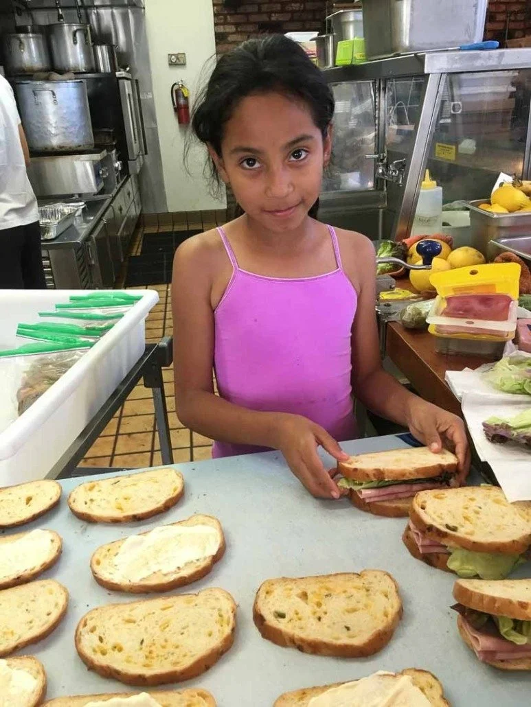A young girl with dark hair in a purple tank top assembling sandwiches with ham, lettuce, and bread behind a table with more sandwiches, in a commercial kitchen.