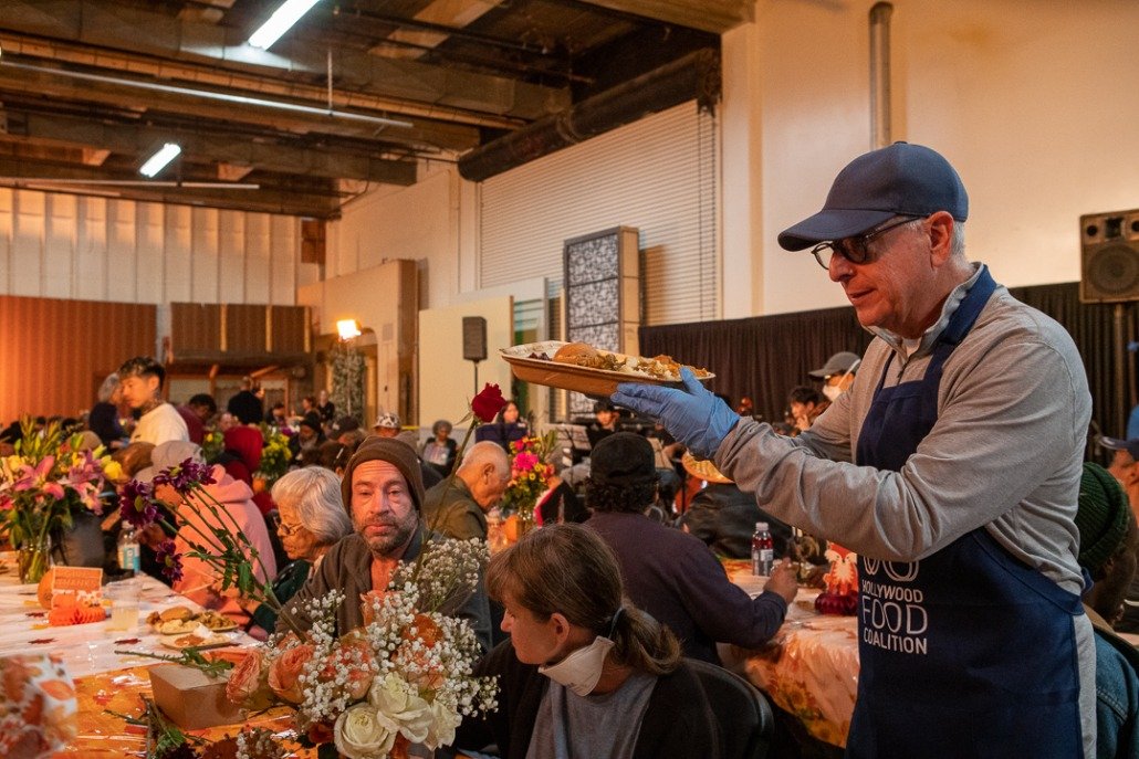 A man with glasses, a cap, and a beard wearing a blue apron serving food at a large indoor gathering with many seated people, flowers, and decorations.