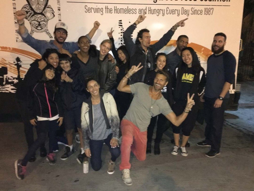 A group of 15 smiling and happy people posing in front of a truck with a banner that reads 'Serving the Homeless and Hungry Every Day Since 1987.' They are making peace and victory signs, some with arms raised, and appear to be celebrating.