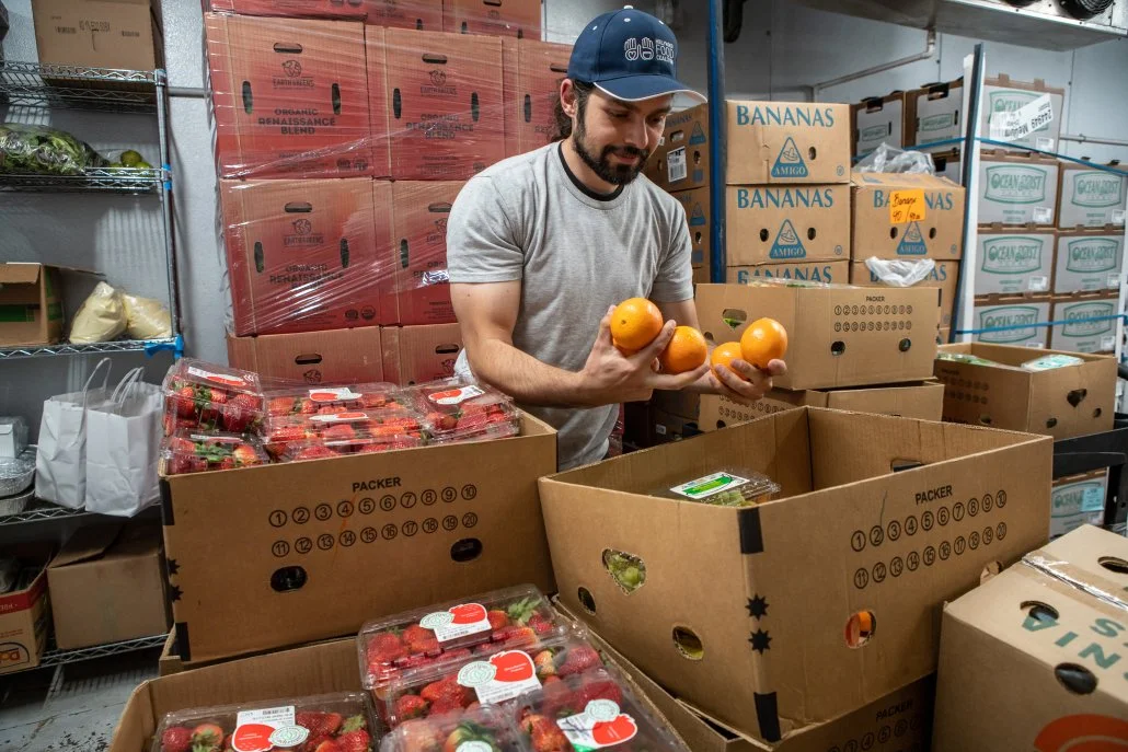 A man in a gray t-shirt and blue cap arranging oranges on a table at a warehouse or storage room, with stacked boxes of produce around him, including strawberries and bananas.