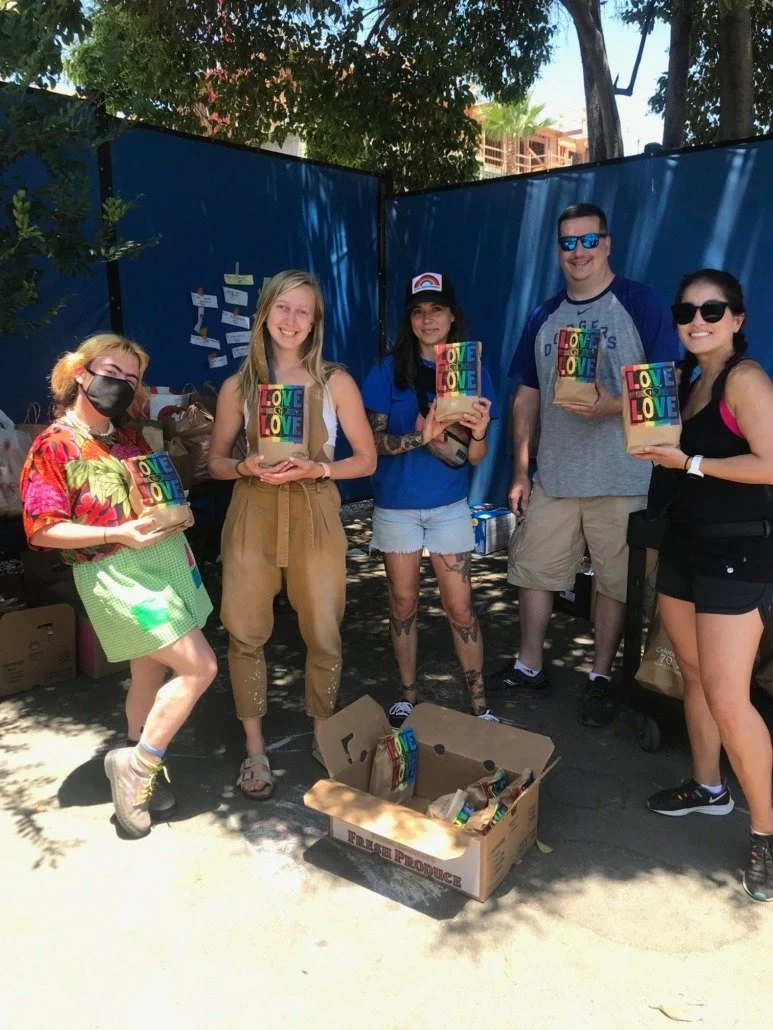 Group of five people standing outdoors, holding colorful gift bags with 'LOVE' written on them, near a cardboard box filled with similar gifts, under shade of trees with construction visible in the background.