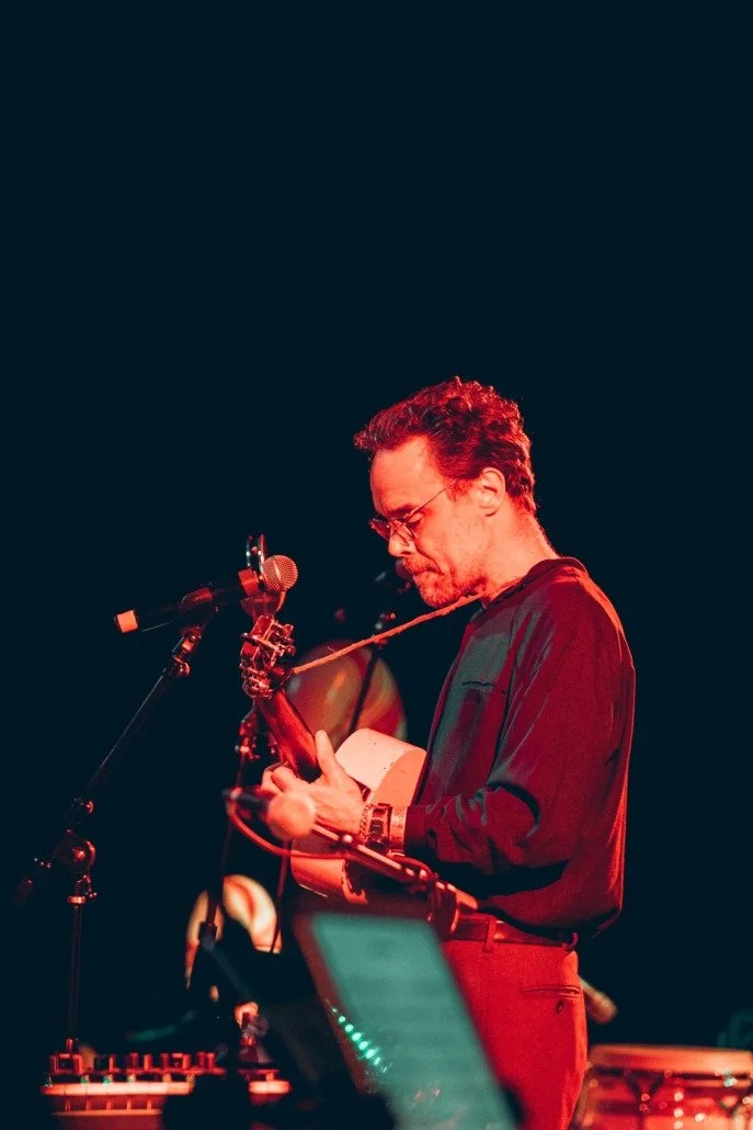 A man playing an acoustic guitar on stage under red lighting, with a microphone in front of him.