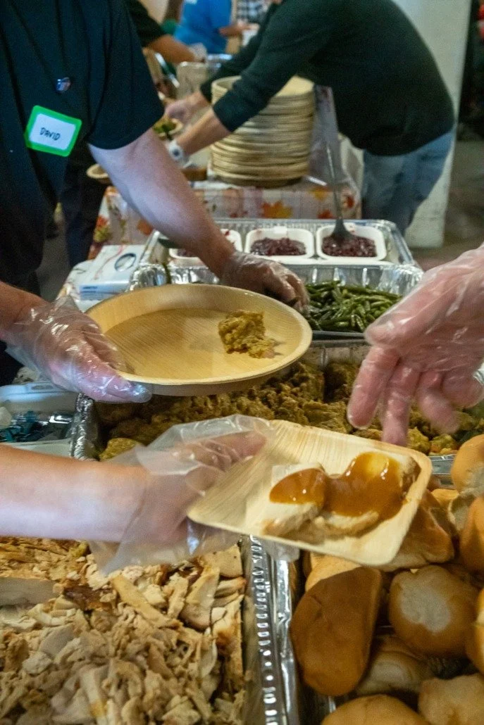 People serving food at a buffet with various dishes including bread, green beans, pulled pork, and baked goods.