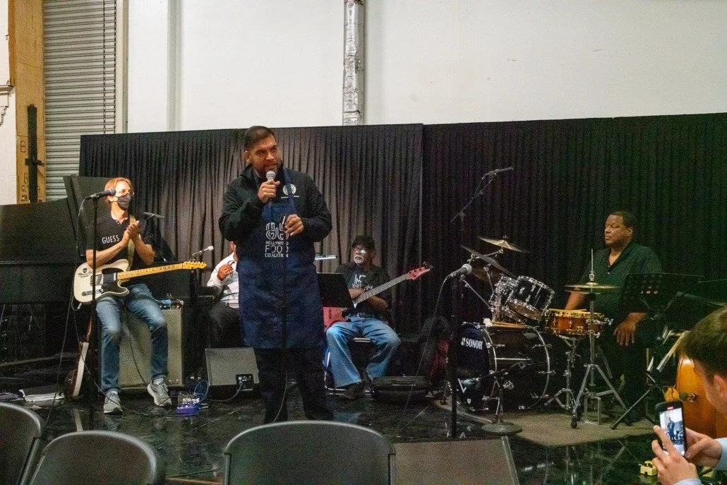 A man holding a microphone and speaking on a stage with a band behind him, including a guitarist, bassist, and drummer, in a room with black curtains.