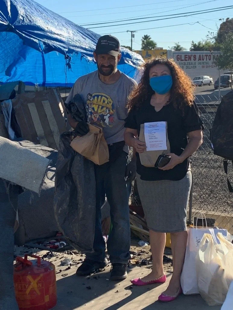 A man and a woman standing outdoors in front of a makeshift tent, wearing face masks. The woman is holding a piece of paper, a mobile phone, and is wearing a grey skirt and pink flats. The man is dressed in casual clothes and a cap that says 'Make Am