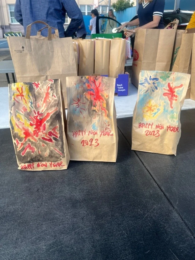 Three paper bags with colorful fireworks and 'Happy New Year 2023' written in red, placed on a black table, with more bags and people in the background at an outdoor event.