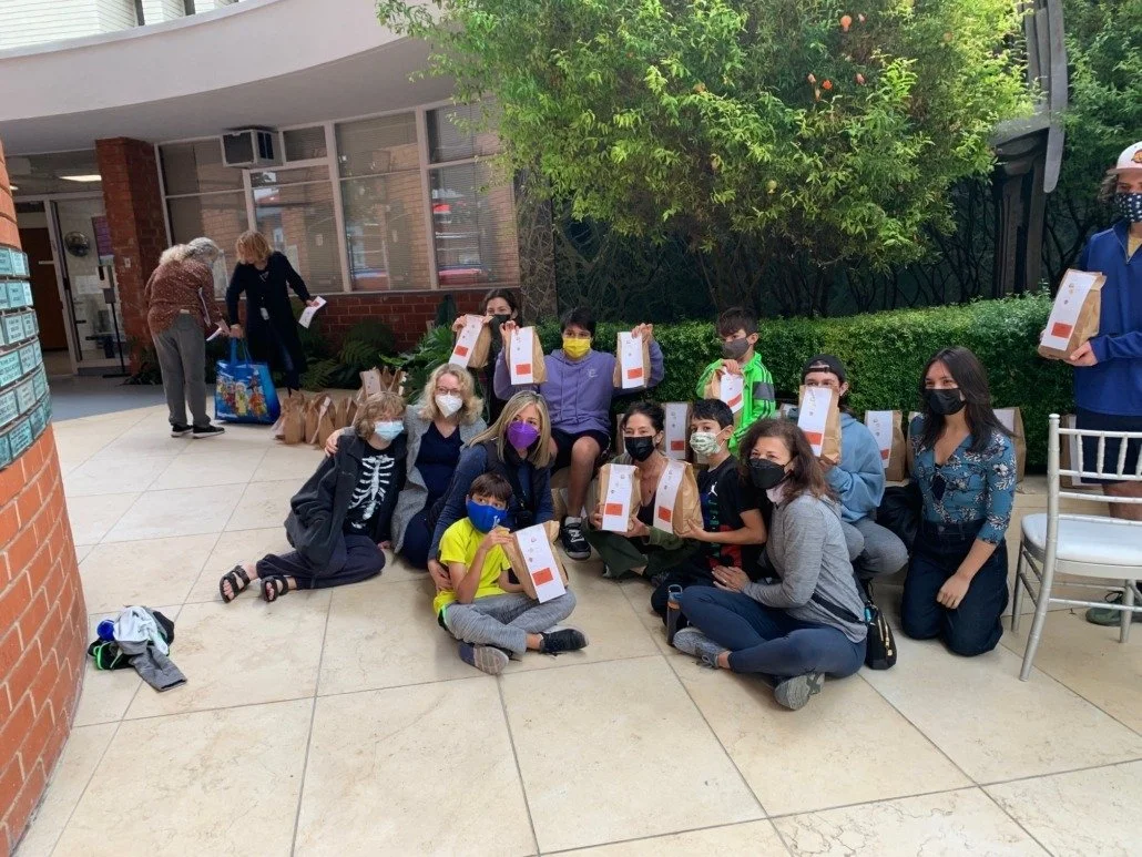 Group of children and adults sitting and kneeling outdoors, holding paper bags, in front of a building with windows and greenery, all wearing masks.