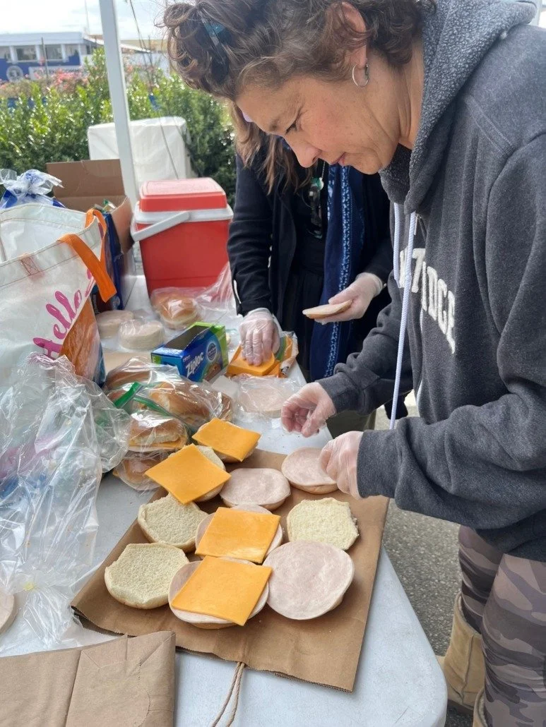 People preparing chicken sandwiches with sliced bread, cheese, and sliced turkey or ham at an outdoor food service table.