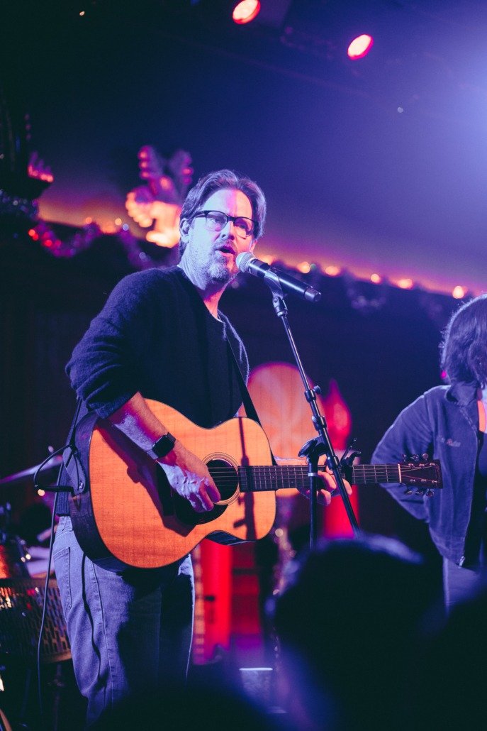 A man with glasses and a beard plays an acoustic guitar and sings into a microphone on stage during a performance at night, with stage lights and a blurred background.