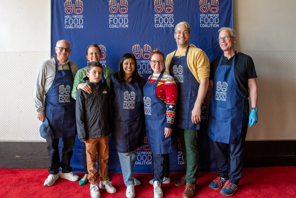 Seven people standing in front of a blue backdrop with the Hollywood Food Coalition logo, wearing aprons. They are smiling and posing for the photo, with a red carpet beneath them.