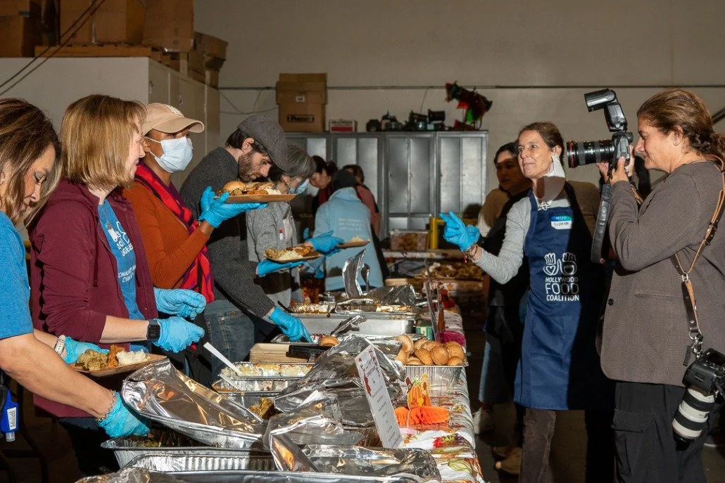 People serving food at a community event, with one person taking photos and others eating or serving behind a table with various dishes.