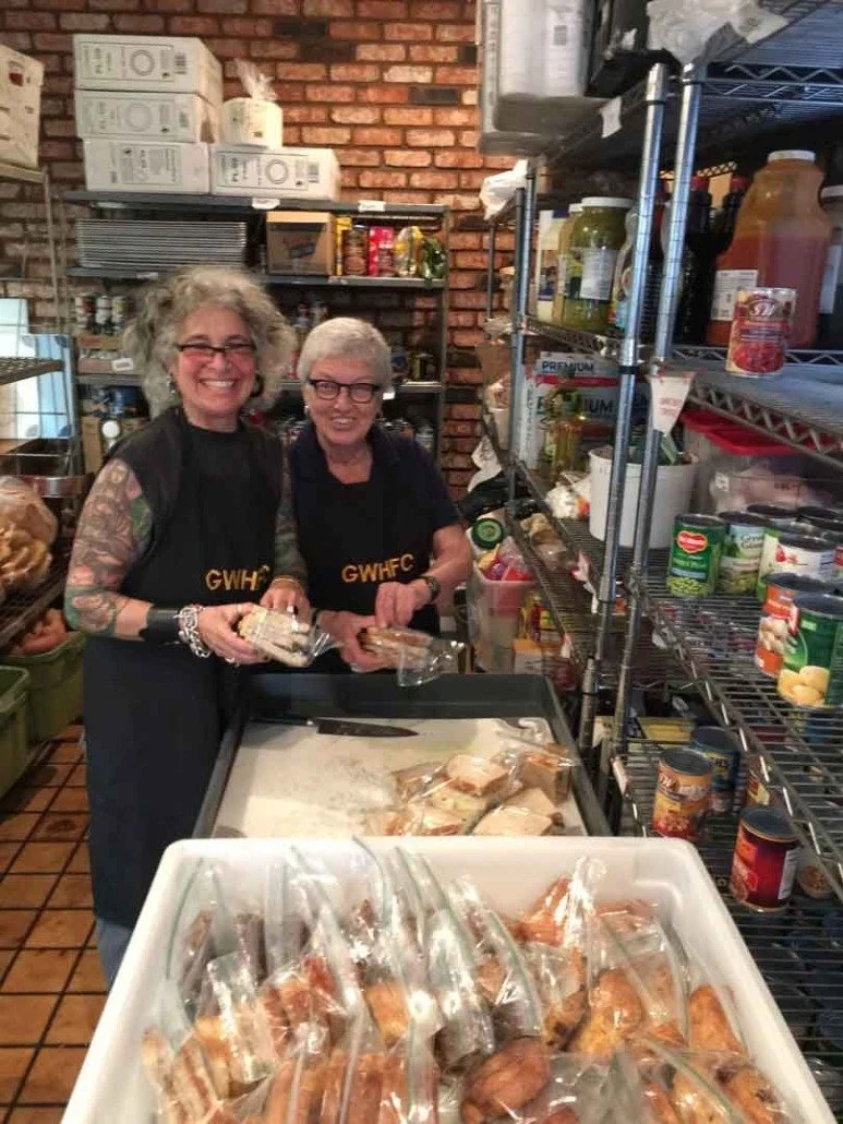 Two women with gray hair and glasses smiling in a kitchen or food pantry. They are wearing black aprons, one with the initials 'GWHFC.' They are packaging sandwiches, with several sealed sandwiches on a tray in front of them and others on a table. Sh