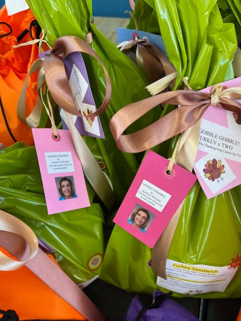 Gift bags with pink and purple tags and ribbon, placed on a surface. The tags feature a photo of Nancy Pelosi and a message thanking her for her service, indicating these are appreciation gifts.