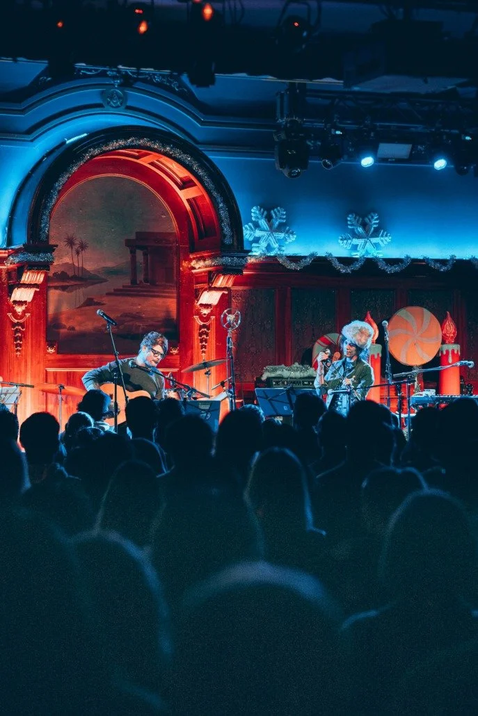 Musicians performing on stage during a holiday-themed concert with Christmas decorations and an audience watching.