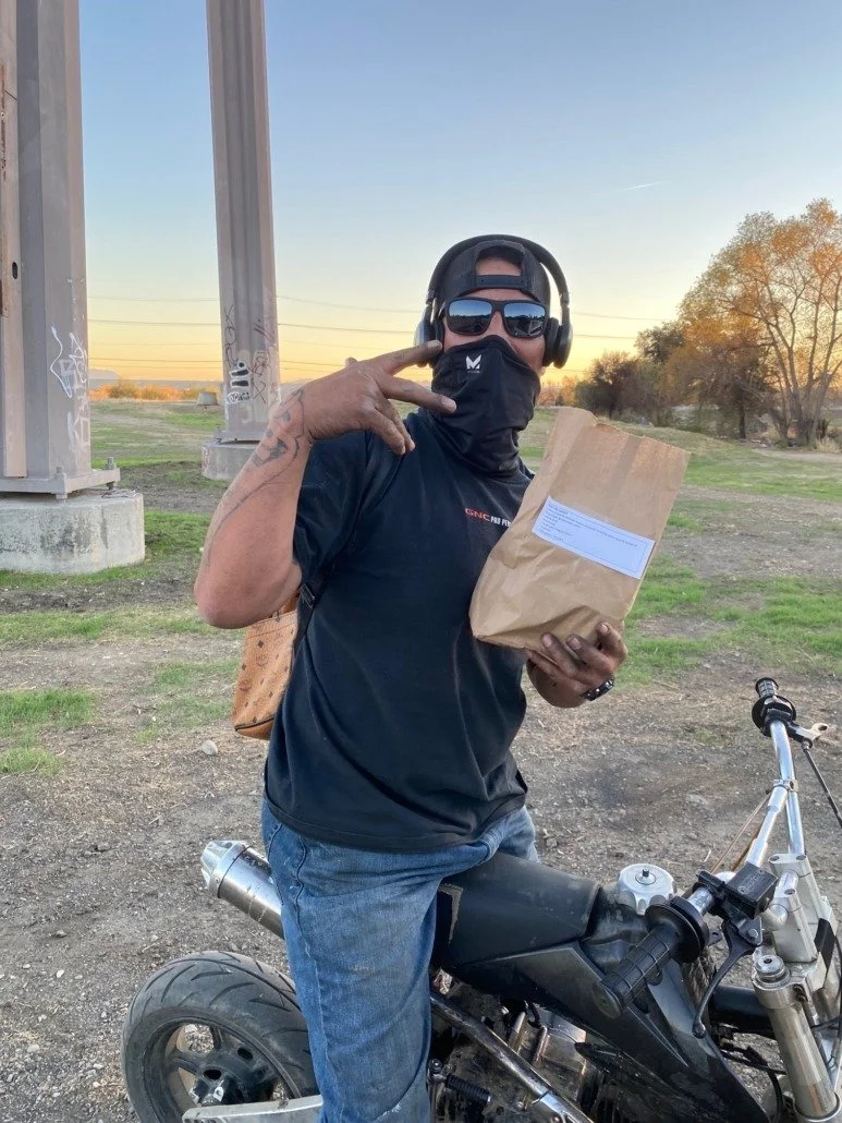 A man wearing a cap, sunglasses, a face mask, a black t-shirt, and jeans standing next to a motorcycle outdoors at sunset, holding a brown paper bag and making a peace sign with his right hand.