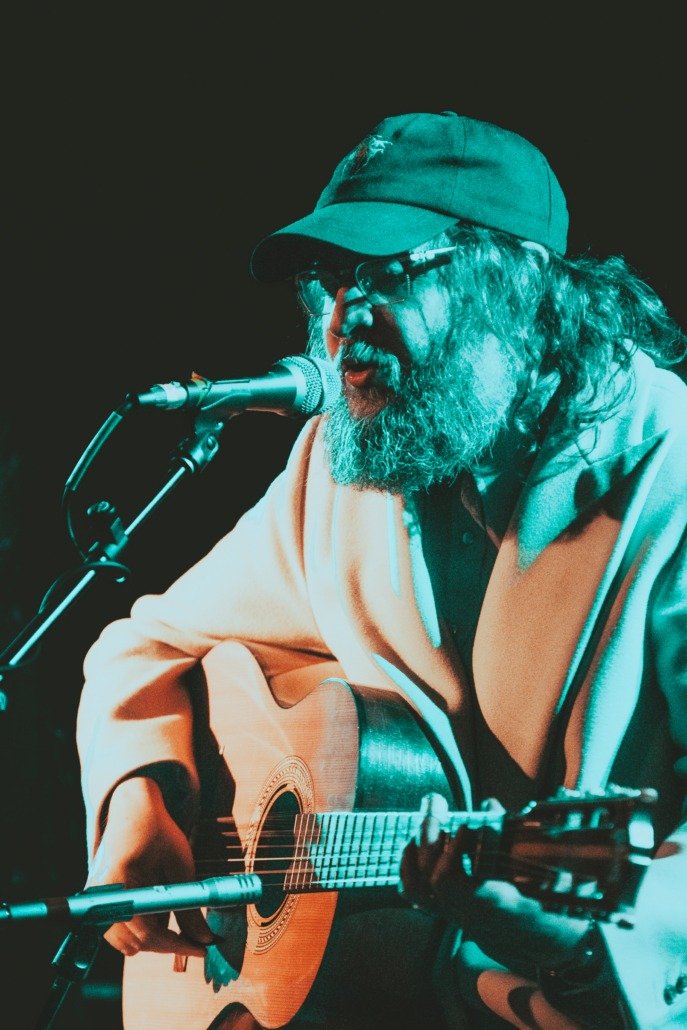 Man with glasses and a beard singing into a microphone while playing an acoustic guitar.