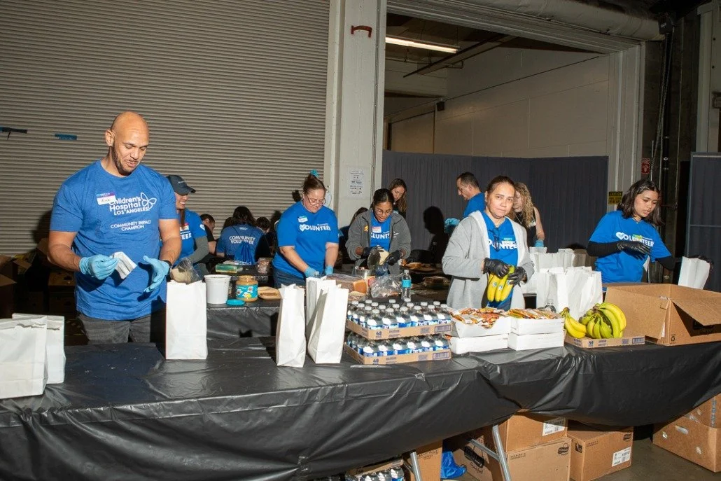Group of volunteers organizing food packages at a community service event in a warehouse, with bananas and bottled water on the table.