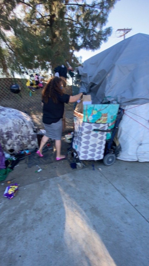 A woman with curly hair, wearing a black top, gray skirt, and pink slippers, is standing outside near a makeshift shelter or tent. She is reaching into a cardboard box, and there is a shopping cart filled with bags and items nearby. The scene is set 