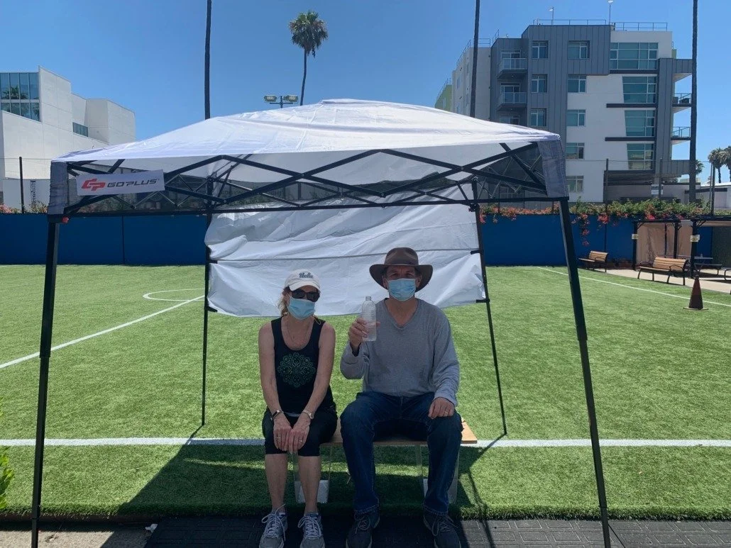 Two people sitting under a canopy on a grassy area, holding water bottles and wearing masks, with buildings and palm trees in the background.