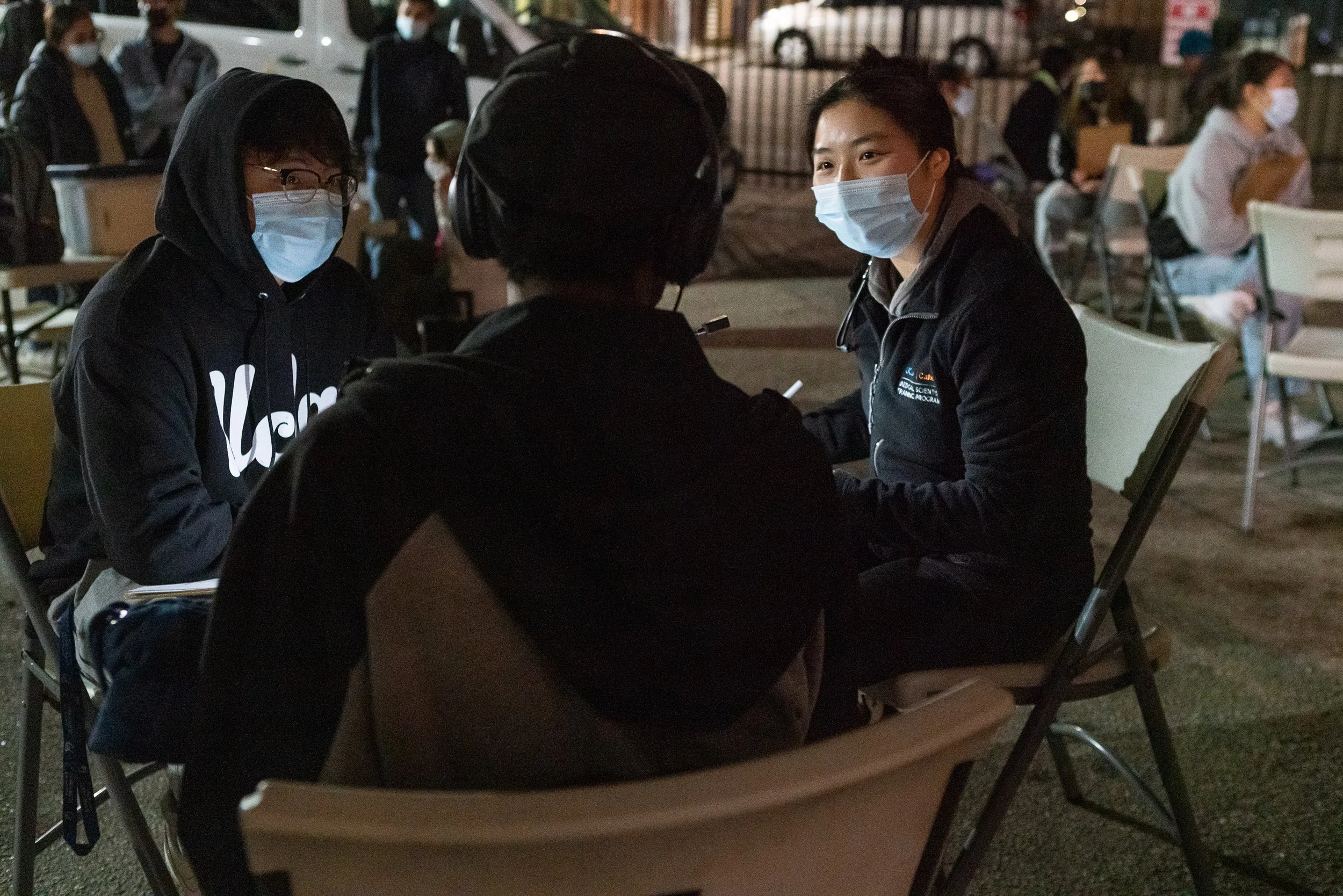 Three people sitting at a table outdoors, wearing masks and engaged in conversation during the evening. In the background, more individuals are seated at other tables, also wearing masks.