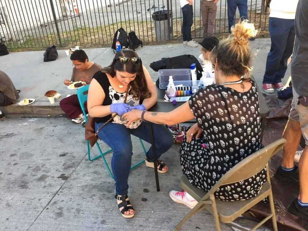 A woman getting a tattoo on her arm at an outdoor event, with several people in the background, some eating and others standing.