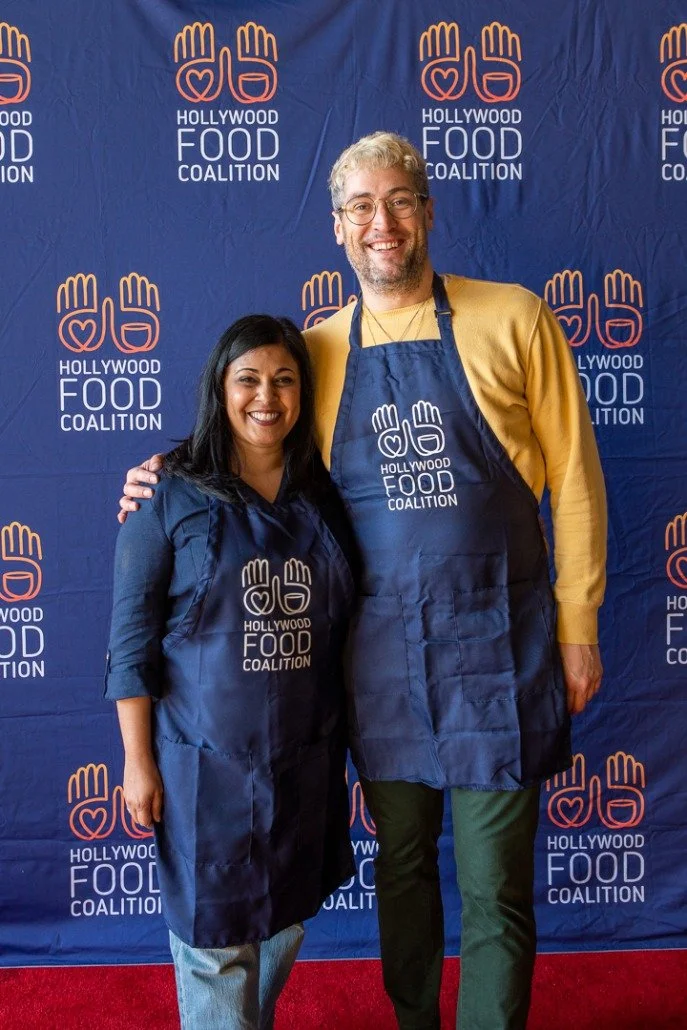 Two people, a woman and a man, standing together wearing blue aprons with the Hollywood Food Coalition logo, in front of a blue backdrop with the same logo, at a charity event.