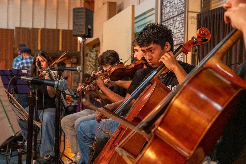 Young musicians playing violins and a cello during a rehearsal or performance in a studio with microphones and music stands.