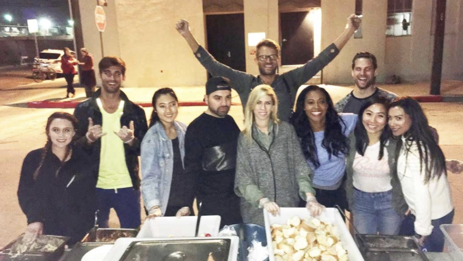 Group of people smiling and posing outside at night, standing behind tables with food, in an urban setting.