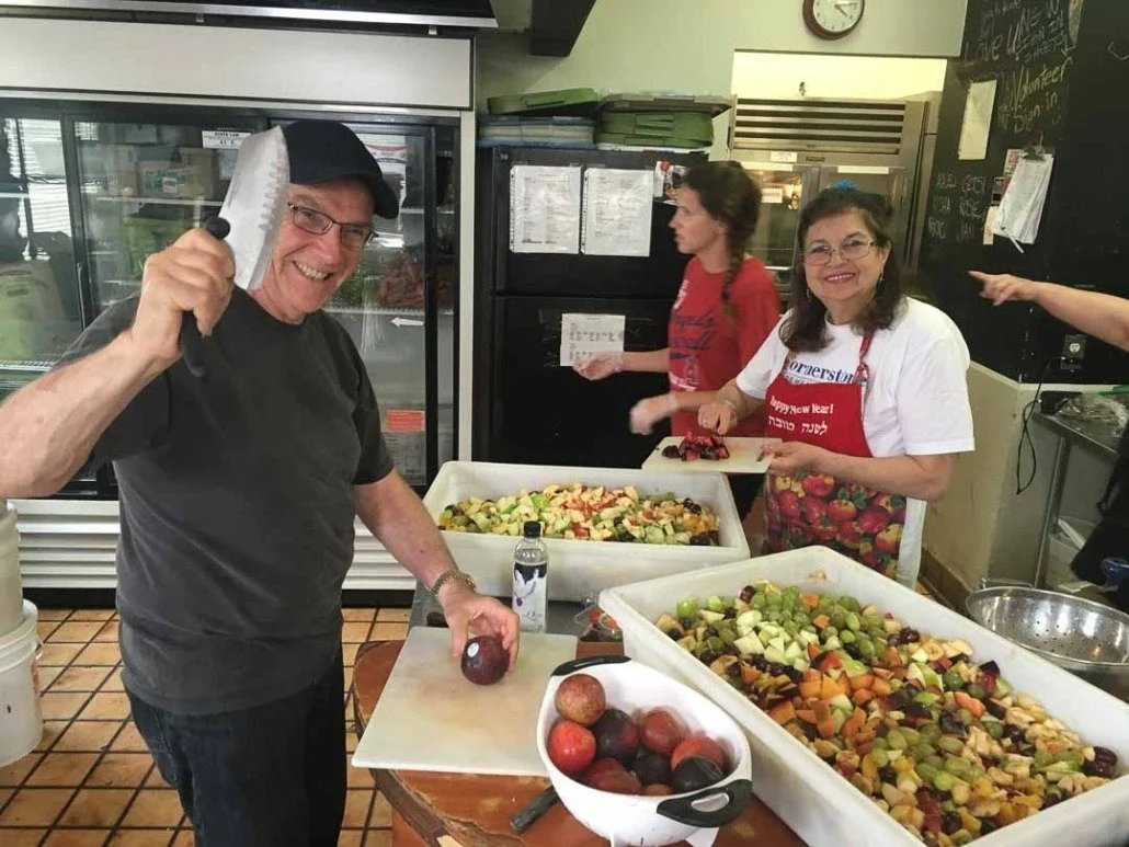 A man holding a large kitchen knife and a red apple in a kitchen with two women preparing fruit salads, one smiling at the camera and wearing a red apron, and the other with a red shirt in the background.