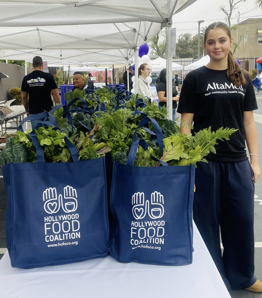 Young woman standing next to two blue bags filled with fresh vegetables at an outdoor event for the Hollywood Food Coalition.